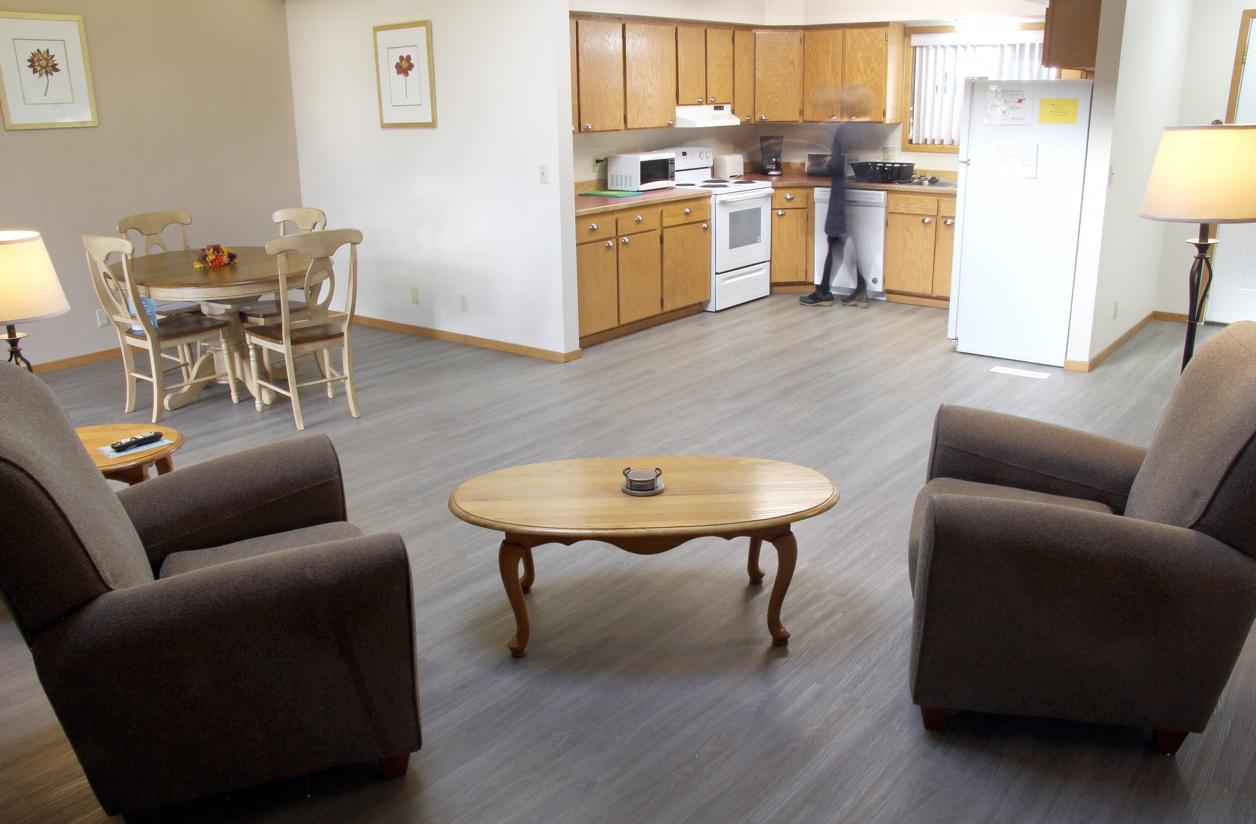 image of the inside of the hugh currin house. In the foreground you can see two armchairs facing each other with a coffee table in between them. on the left side of the room there is a round dining table that seats four people. the right side of the background reveals a full kitchen with a stove, microwave,dishwasher, and refrigerator. The Hugh Currin house is available for patients receiving cancer treatment in klamath falls and need a place to stay. it is across the street from the cancer treatment center.