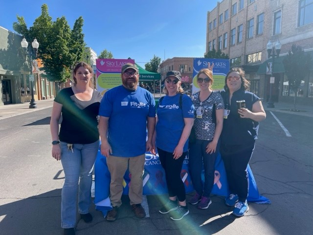 cancer treatment center staff standing in front of a cancer treatment center booth at a summer third thursday event in downtown klamath falls. the sun is bright. there are five people in the image: a tall woman on the far left wearing a black tee-shirt and jeans. a man with a beard in a ball cap wearing a blue tee shirt that says "fit 2 be cancer free" and khakis. another woman wearing sunglasses and a ball cap wearing a blue fit 2 be cancer free tee shirt and dark pants. Another woman wearing sunglasses and scrubs, she had a Sky Lakes staff badge that says "R.N' the woman next to her is holding bunny ear fingers behind her head. Ont he far right a woman wearing sunglasses with dark hair is wearing dark clothes and has a sky lakes staff badge and holding a beverage.