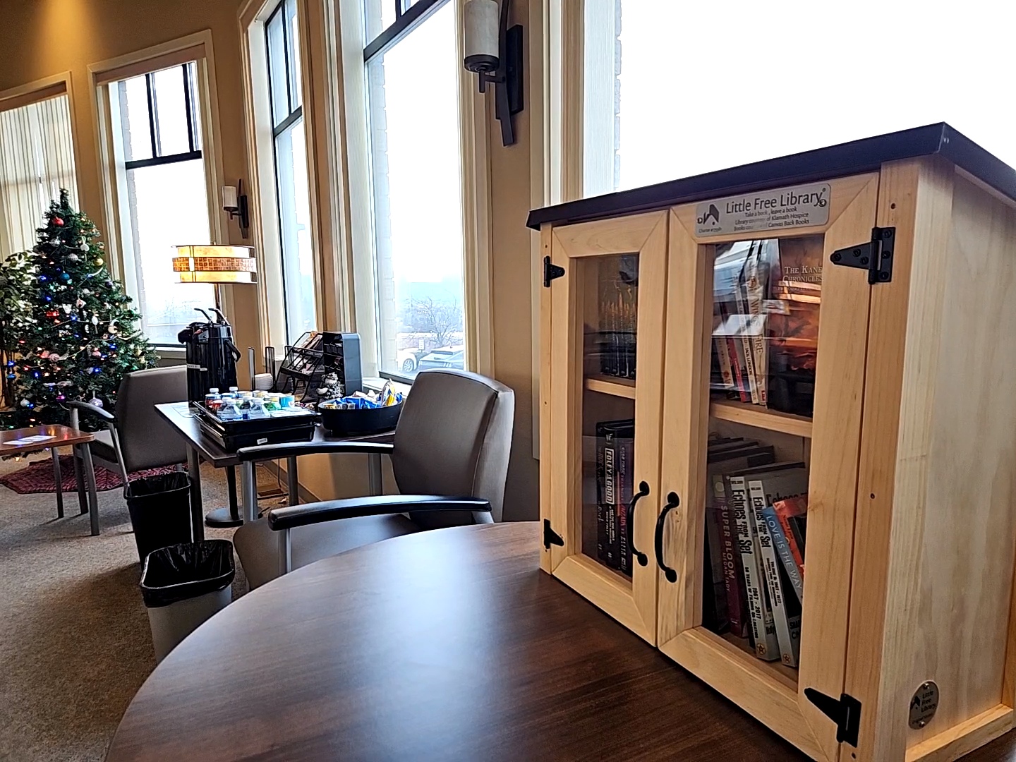 image of a little free library full of books sitting on a brown round table in the sky lakes cancer treatment center in the foreground. In the background there are tables with refreshments for patients and a decorated christmas tree. There is bright natural light coming through the windows.