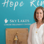 Nurse Hannah Sonerholm standing in front of a blue wall that says "let hope Ring' and "Sky Lakes Cancer Treatment Center"