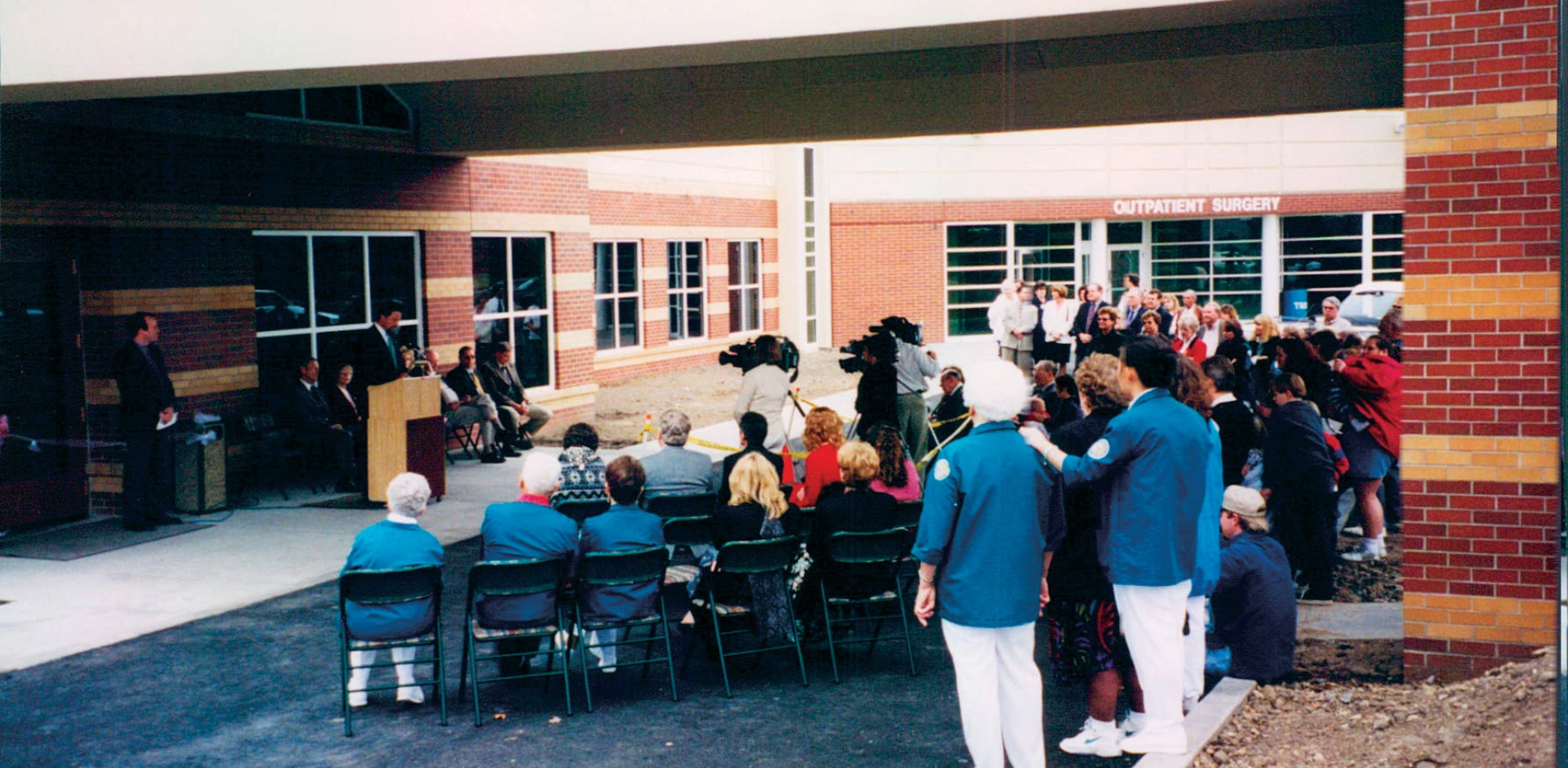 The Opening Ceremony for the Family Birth Center. there's a group of people outside the main doors. A man addresses the crowd at a podium. Other people stand behind him.