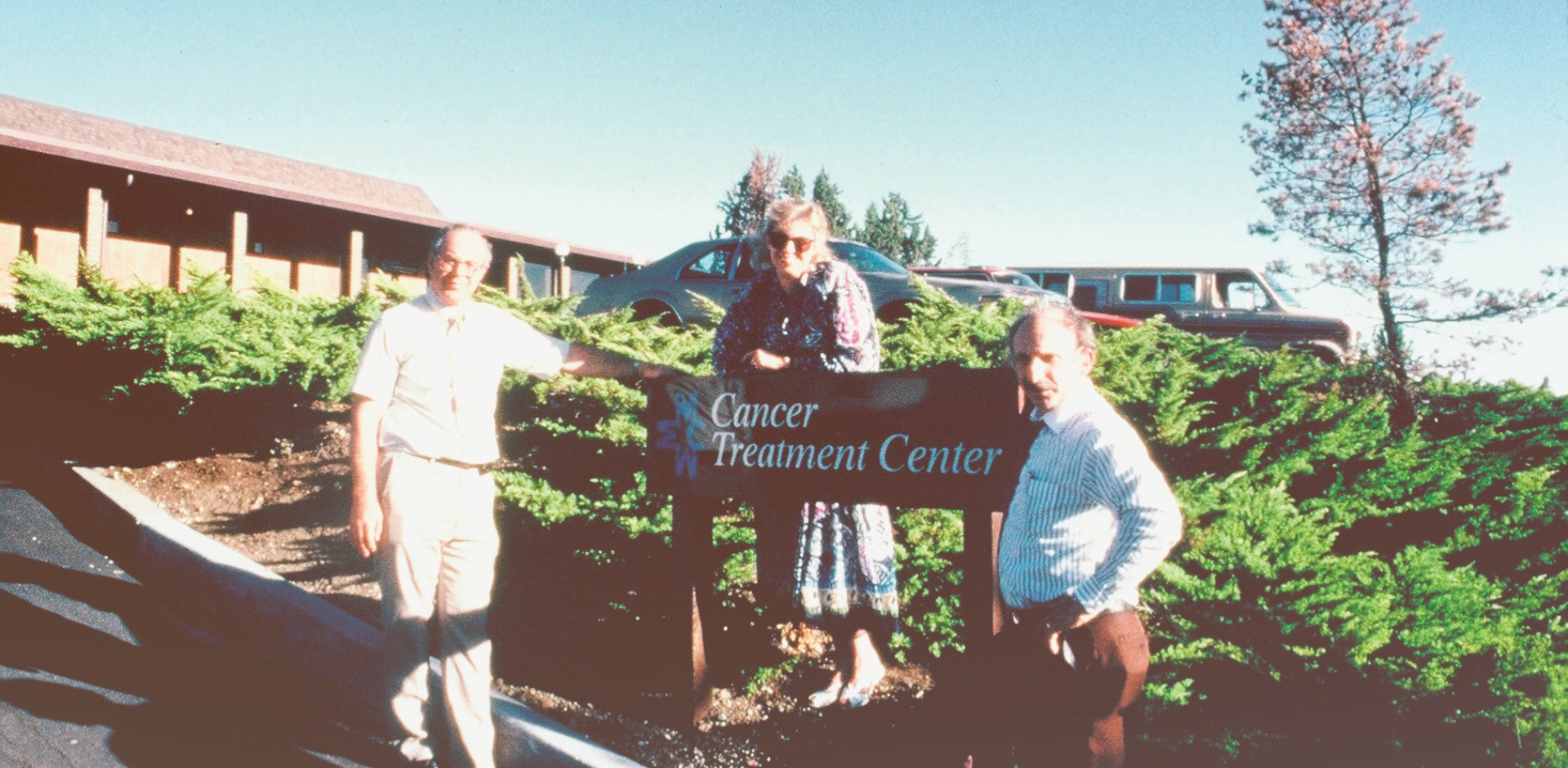 An old photo of three people standing outside the Merle West cancer treatment center in front of the sign for the building. Taken in the late 1980s.