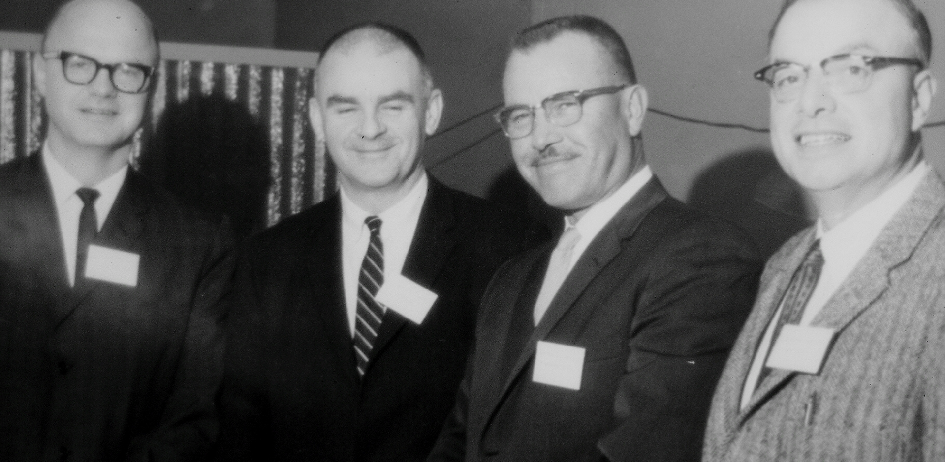 Four men smile at camera, photo is in black and white. t/aken in the 1960s.